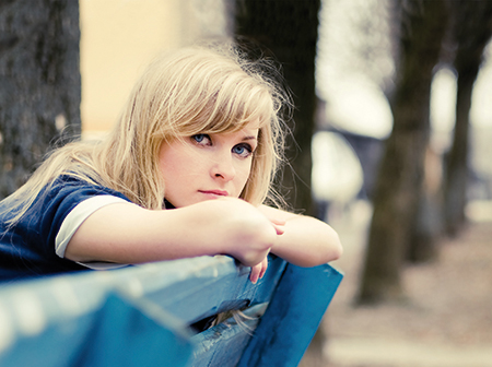 Eine junge Frau sitzt auf einer Parkbank und stützt ihren Kopf auf die Unterarme, die auf der Rückenlehne liegen. 